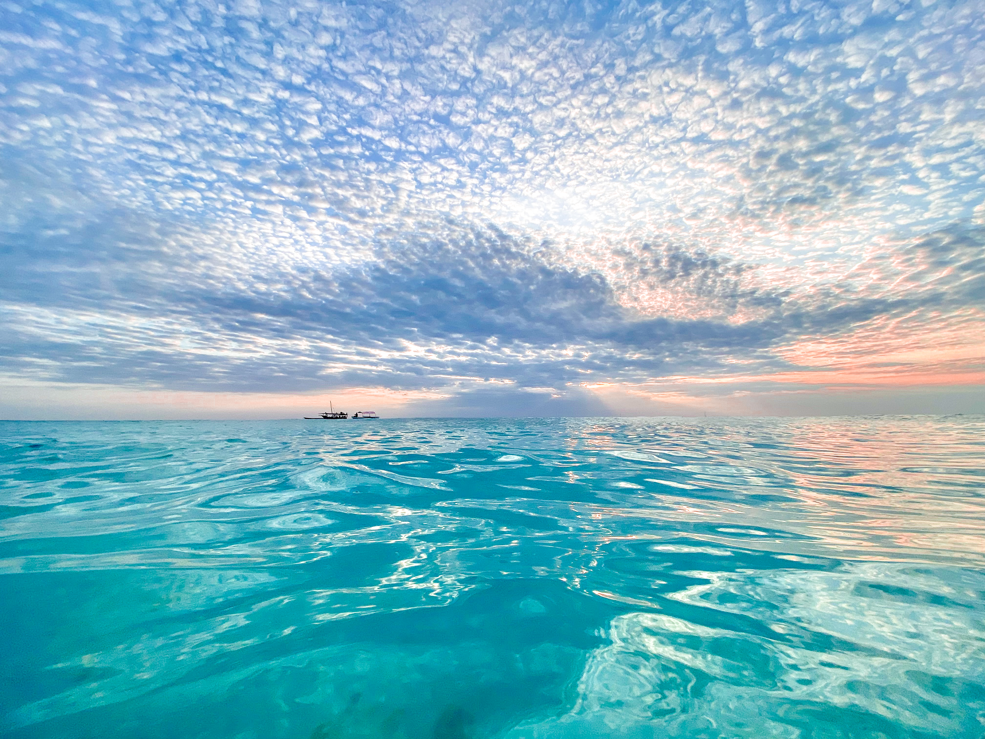 Clear turquoise ocean water in Nungwi Beach, Zanzibar at sunset with small boats on the horizon under a textured cloudy sky.